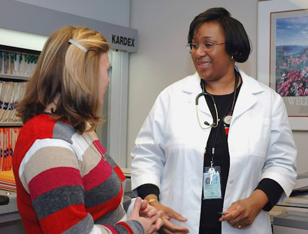 A smiling female doctor wearing a stethoscope and white coat engages in a conversation with a female patient wearing a striped sweater in a medical office.