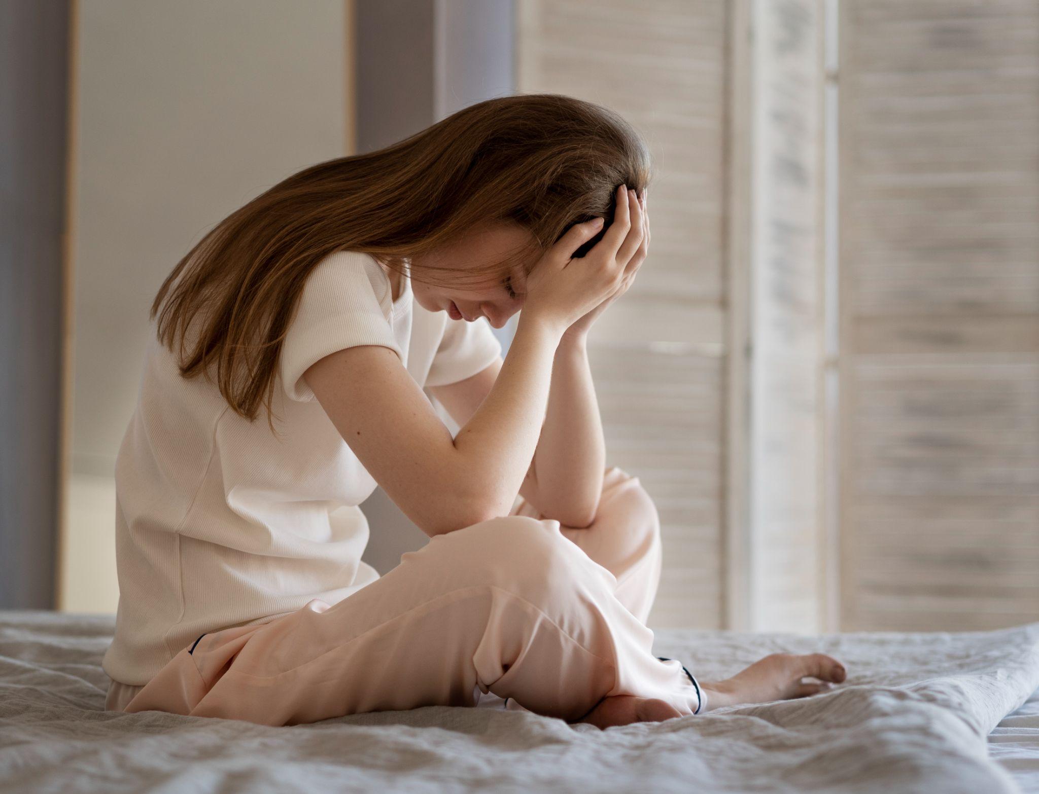 A woman sitting cross-legged on a bed in pajamas, holding her head in her hands with her face hidden in a gesture of distress or pain.